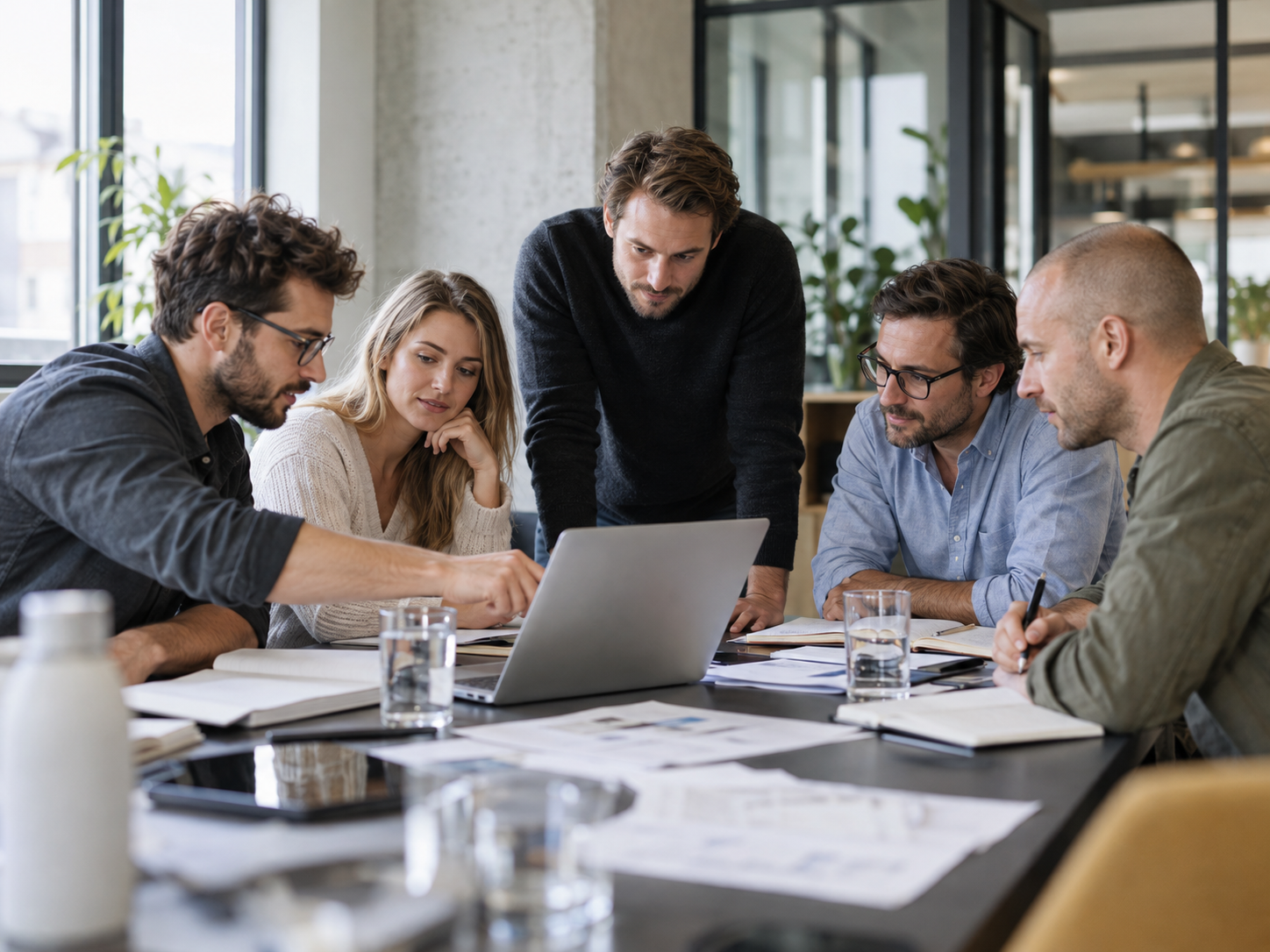 Fünf Teammitglieder arbeiten gemeinsam am Laptop und besprechen Unterlagen an einem Arbeitstisch. KI-generierte Abbildung.